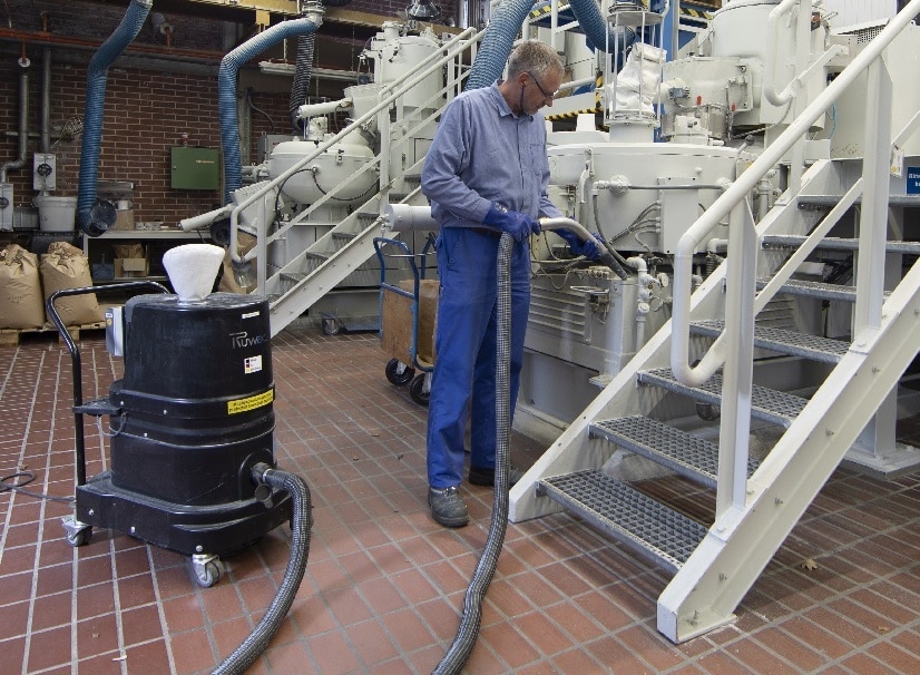 Mitarbeiter einer Compoundieranlage saugt zwischen zwei Industrietreppen Staub von den Geräten.