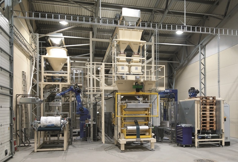 In einer Industriehalle stehen zwei Verpackungsstationen. Die linke wird durch einen Roboter bedient.