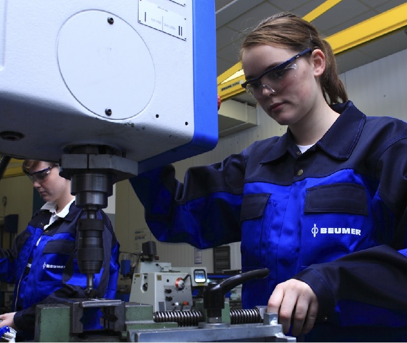 Junge Frau steht an einer mechanischen Apparatur des Maschinenbaus. Im Hintergrund links ein anderer Lehrling.