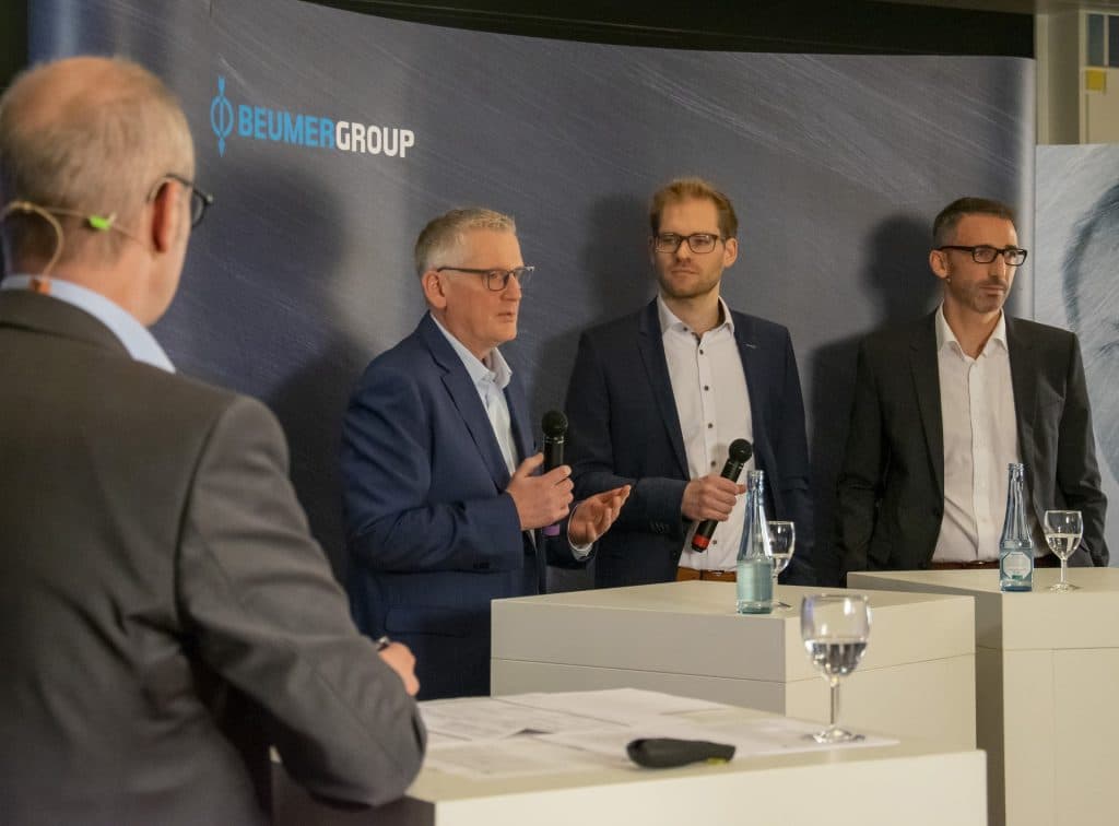 Intervieview Situation, 3 Gäste die man sieht und ein Moderator den man nur von hinten sieht, vor einer BEUMER GROUP Messewand.