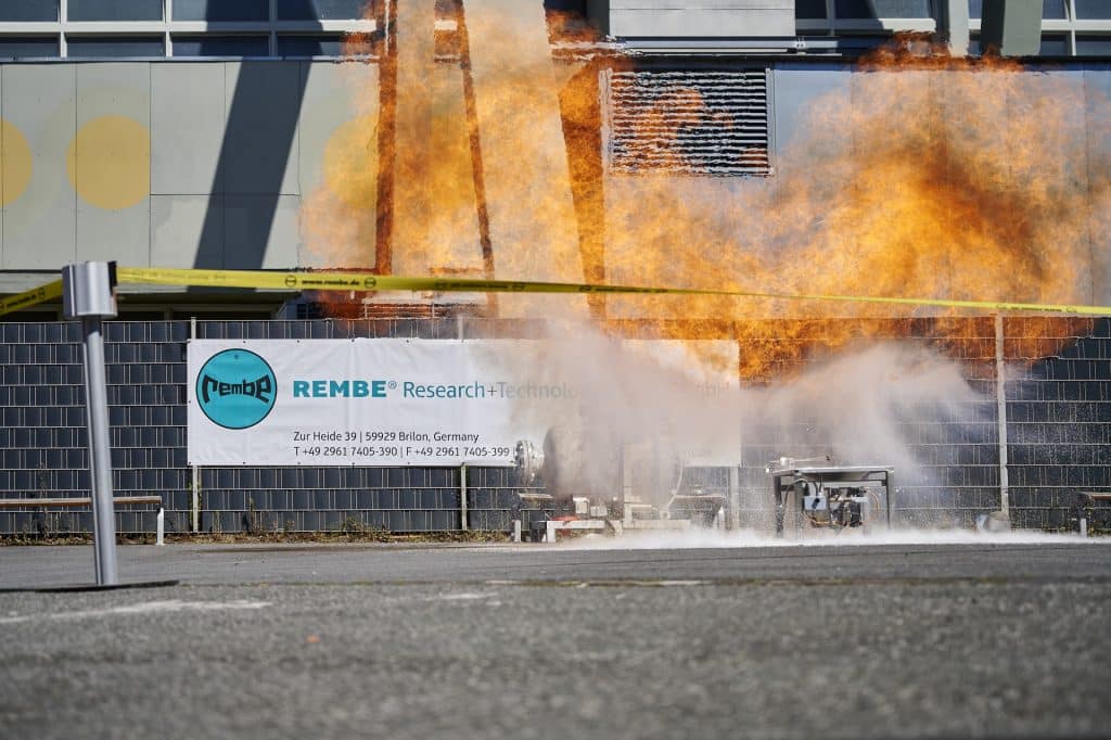 Life explosions Show der REMBE® GmbH Safety + Control auf der Solids in Dortmund. Im Hintergrund die Messehalle und ein Banner der Rembe Research. Im Vordergrund oben Flammen, unten Rauch darunter der Veruchsaufbau. Auf schuettgutmagazin.de