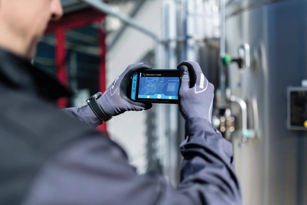 Seitenansicht eines Mitarbeiters vor einer Industrieanlage mit einem Smartphone in der Hand, auf schuettgutmagazin.de