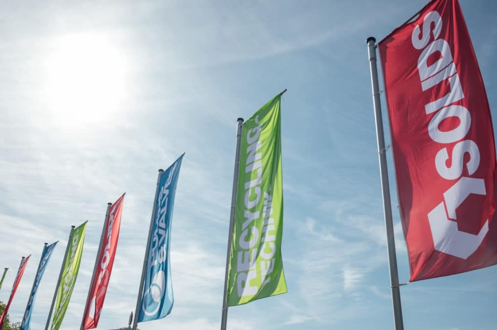 Viele Fahnen vor blauem Himmel, mit alternierend roter Fahne für die Messe Solids Dortmund, grün für die Recycling-Technik, und blau für die Pumps & Valves