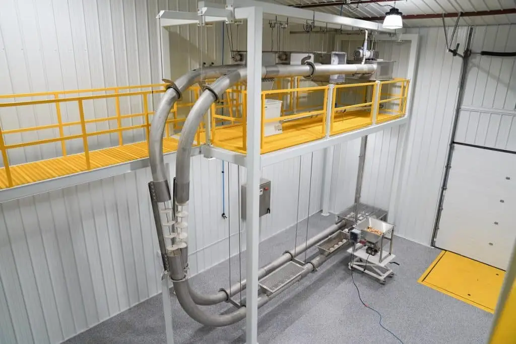 Industrie-Bau in einer Halle: Materialtransportsystem, das mit Schläuchen/Rohren über zwei Stockwerke arbeitet. Gelbe mit Begrenzungen geschützte Gehwege im Obergeschoss.