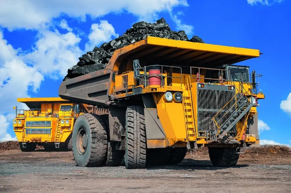 Zwei Baustellen, Schwerlaster in gelber Farbe auf einem Außengelände. Steinkohle ist bei dem ersten Laster als Ladung sichtbar. Blauer Himmel mit hellen Wolken.