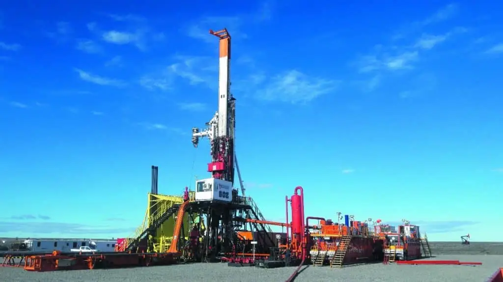 Tiefbohranlage im Vordergrund. Links im Hintergrund eine Pumpe, vielleicht Öl. Industrieanlage mit einem turmartigen Komplex. Rechts davor Tankanlagen.