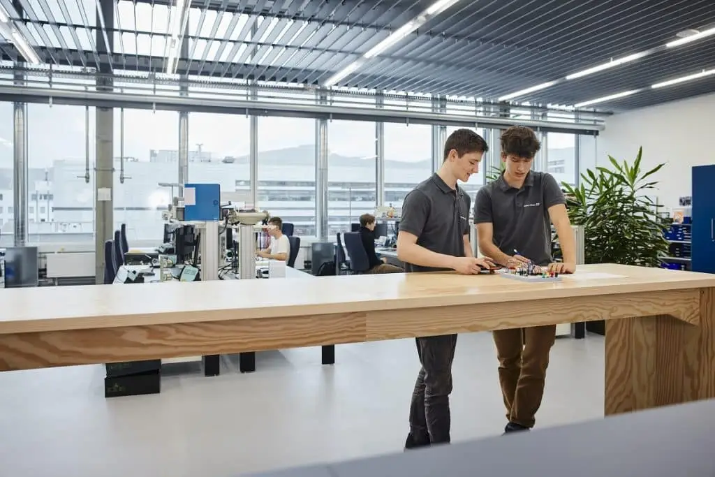 Ein Büro bei Endress+Hauser. Zwei junge Männer stehen an einem langen Holztisch und schauen sich scheinbar ein Schaltmodell an. Im Hintergrund sitzten andere junge Menschen an Arbeitsplätzen für Auszubildende.