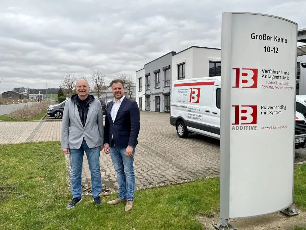 Vor dem Firmengelände links vom Firmenaufsteller stehen Frank Scholz, Inhaber und Geschäftsführer der IB Verfahrens- und Anlagentechnik GmbH & Co. KG. (links) und Floris Rouw, Leiter des Geschäftsbereichs Vakuumfördertechnik von Piab (rechts) auf einem kleinen Stück Rasen. Rechts von Ihnen ein Firmenbus mit Logo und Text der IB Verfahrenstechnik, rechts davon das zweistöckige Firmengebäude.