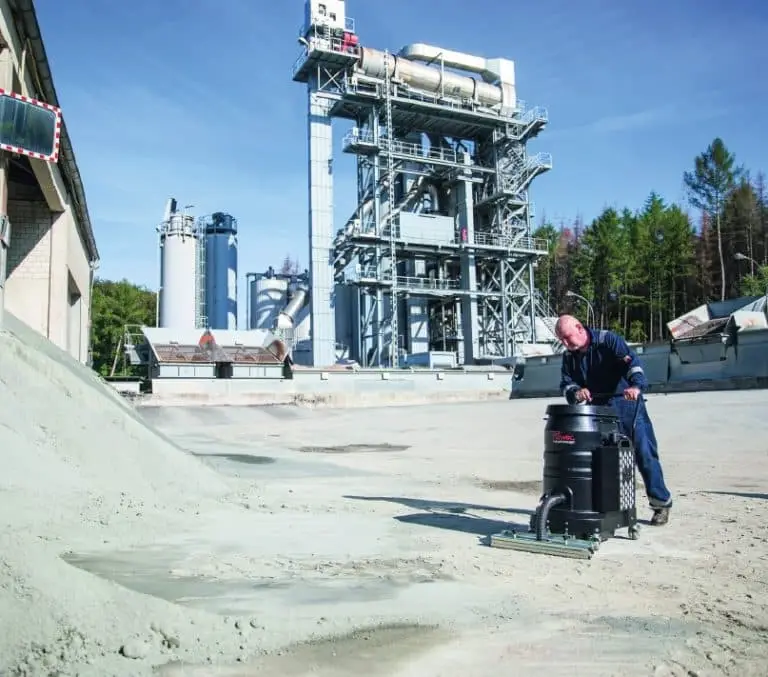 Sandiger Außenbereich eines Industriehofs. Vorne rechts im Bild ein Mann, der mit einem Industriesauger feine Erde einsaugt.