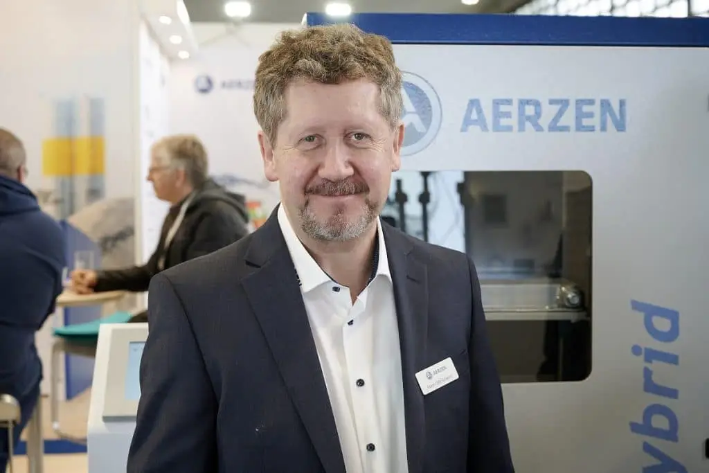 Horst Ulirk Schwarz der Aerzener Maschinenfabrik steht vor einem Ausstellungsstück auf dem Solids 2023 Messestand. Links hinter ihm zwei Messebesucher an einem Tisch im Gespräch.