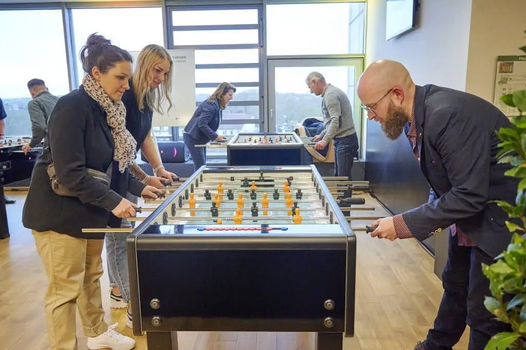 Drei Personen stehen an dem vorderen Kicker und spielen gegeneinander, dahinter zwei Spieler an einem anderen Kicker. Links im Hintergrund ein weiterer Kicker mit Spielern.