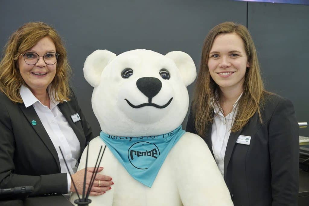 Ute Weber und Lea Friedewald mit dem Rembe Stoffeisbären, der ein Dreieckstuch mit dem Rembe Logo um den Hals trägt auf dem Messestand des Unternehmens auf der Solids 2023
