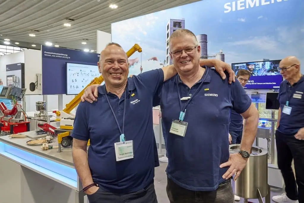Holger Bohlmann und Christoph Hildebrand - von links nach rechts auf ihrem Siemens Messestand der Solids 2023. Im Hintergrund Ausstellungsstücke und Kollegen sowie ein Bildschirm