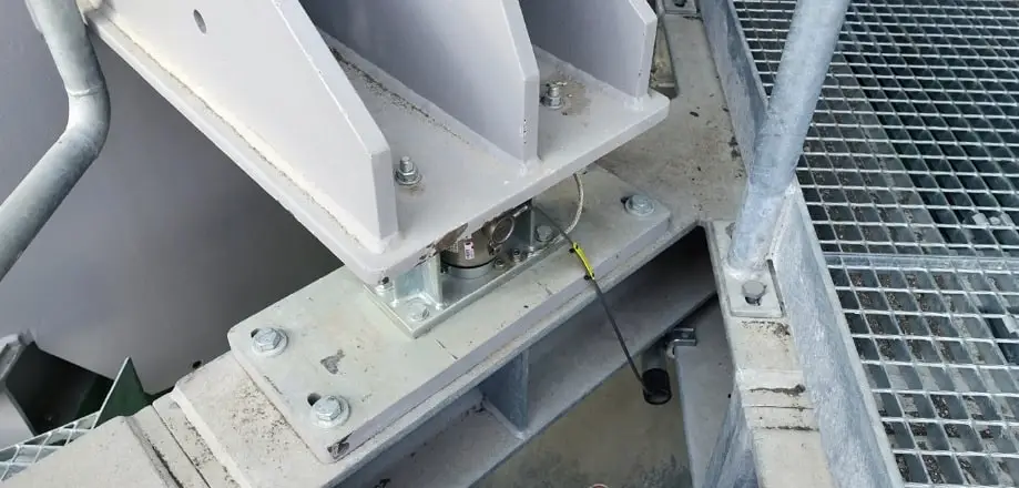 Wägezelle zwischen zwei Stahlträgern in einem Industriegebäude. Ein Draht führt von der Wägezelle nach unten in ein Rohr. Rechts davon ein begehbares Gitter.