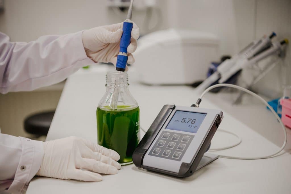 Laborsituation: zwei Hände messen mit einem Endress+Hauser Messstab in einer Laborflasche eine grüne Flüssigkeit auf ph-Wert und Temperatur. Das Messgerät steht rechts von der Flasche.