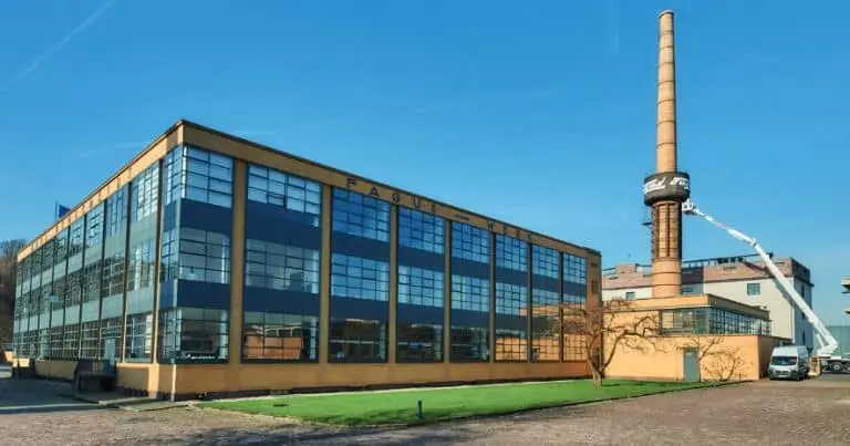 Bauhaus Industriegebäude mit viel Fenster. Rechts davon noch ein kleineres Gebäude mit einem Schornstein. Dieser wird gerade von einem Menschen auf einem Hubwagen besucht.