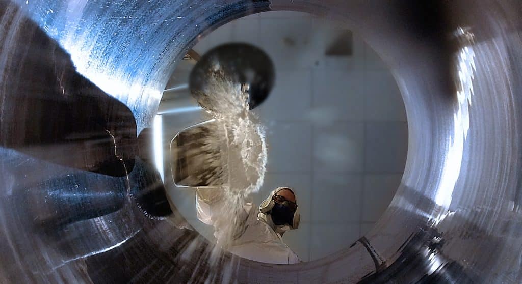 Ein Mitarbeiter der Stasskol füllt mit Atemmaske und in weißer Schutzkleidung ein weißes Pulver in einen Behälter. Das Bild ist aus der Bodenperspektive des Behälters fotografiert.