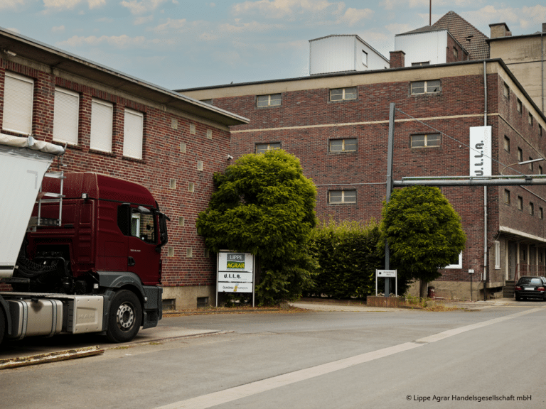 Links ein Ausschnitt eines Lastwagens, der auf einer Bodenwaage steht. Das Ganze ist auf dem Hof eines Getreidehandels fotografiert. Die Häuser sind aus dunklem Ziegelstein.