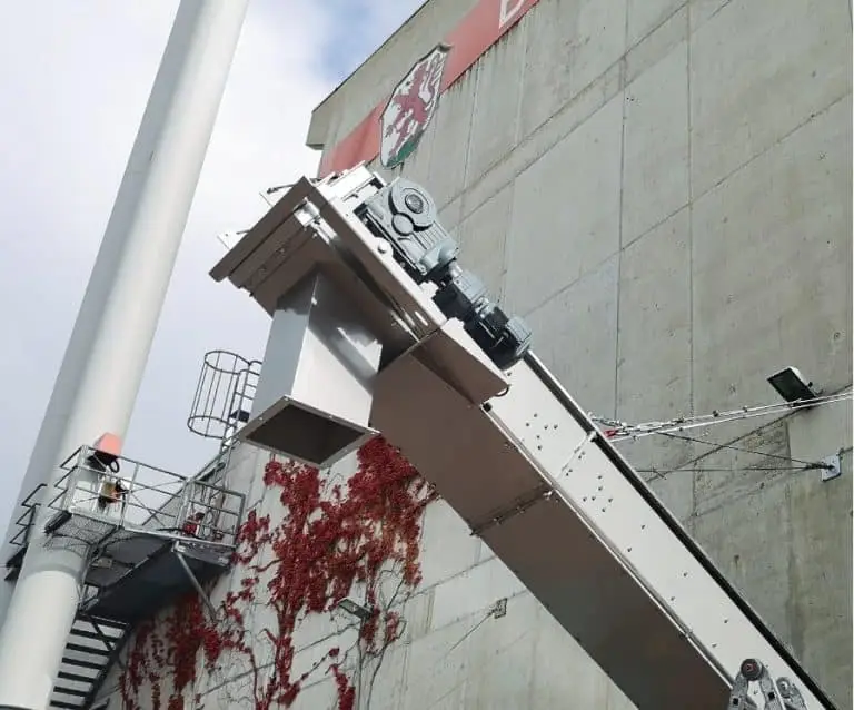 Industriehalle von Außen mit einem Schornstein links und der grauen Betonwand, die teilweise mit rotem Laub bewachsen ist im Hintergrund. Rechts im Vordergrund ein aus der Halle ragendes umhaustes Förderband.
