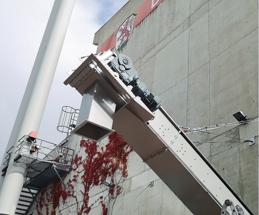 Industriehalle von Außen mit einem Schornstein links und der grauen Betonwand, die teilweise mit rotem Laub bewachsen ist im Hintergrund. Rechts im Vordergrund ein aus der Halle ragendes umhaustes Förderband.
