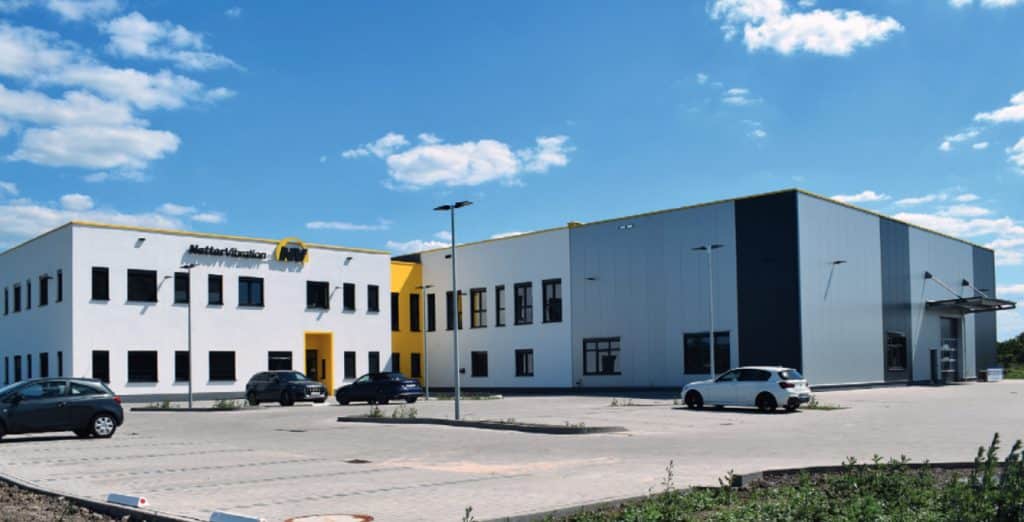 Firmengebäude mit Produktionshalle in Industriegebiet über den Parkplatz hinweg. Gelbe Farbelemente und das Logo der Netter Vibration sind zu sehen.