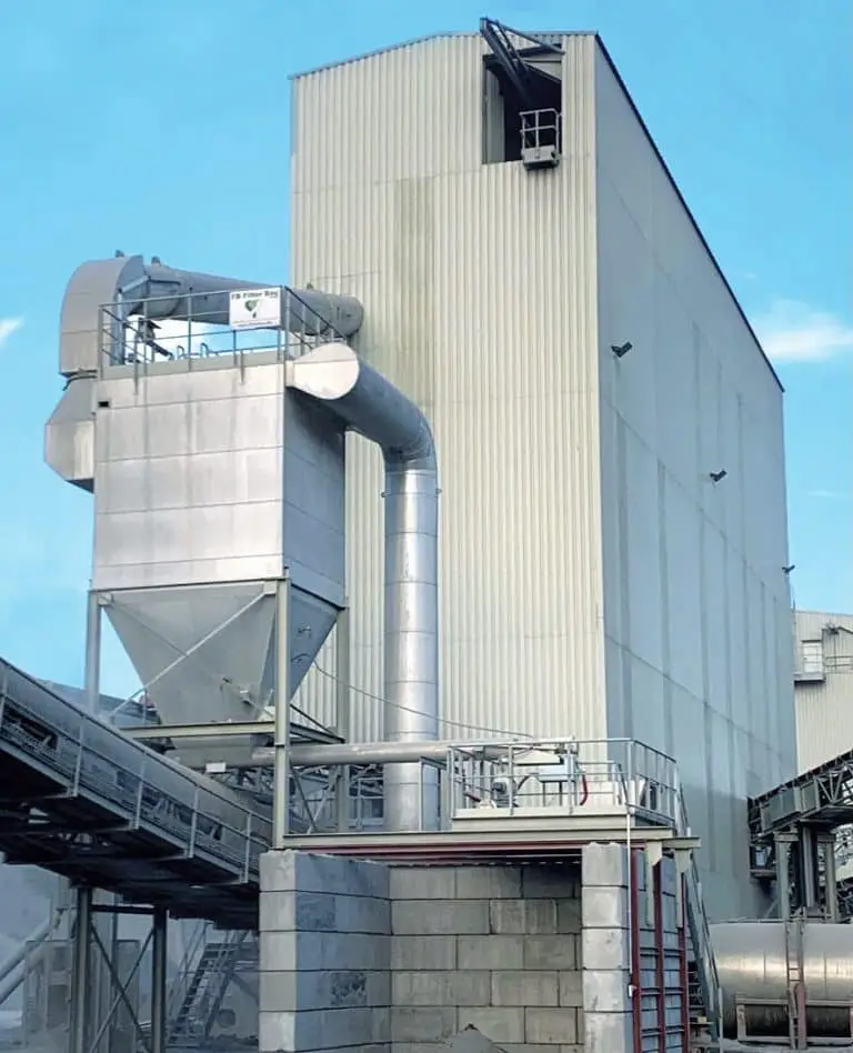 Entstaubungsanlage vor einem Industriegebäude bei blauen Himmel. Vorne ein überdachtes Förderband, bei der Übergabestelle darüber die Entstaubungsanlage beim Dosiersilo. Im Hintergrund eine Hohe Lagerhalle