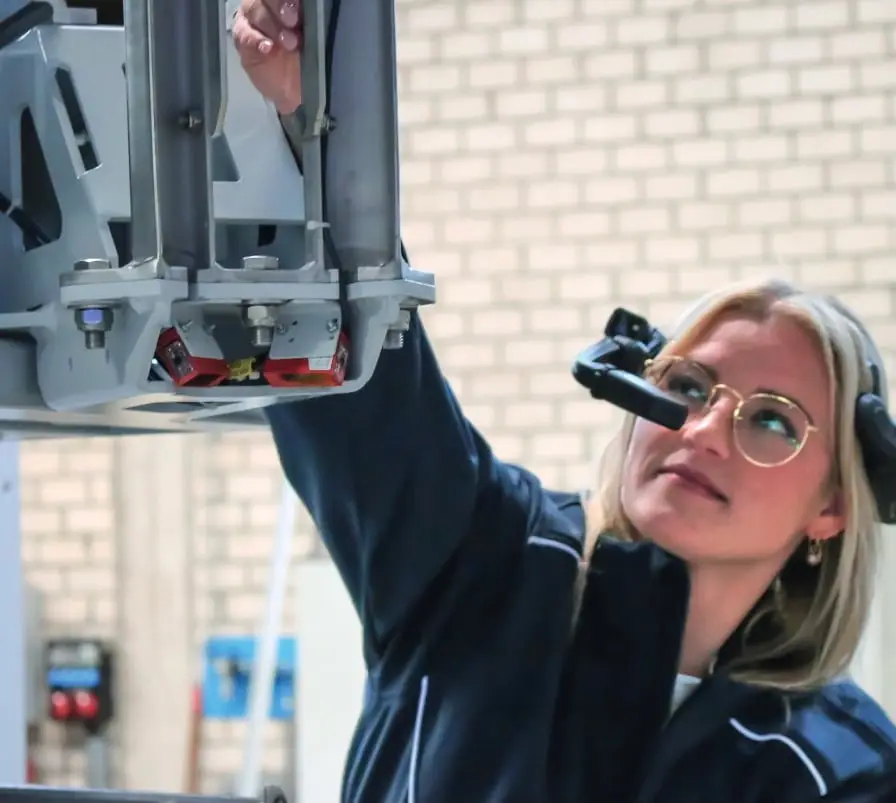 Rechts eine junge Frau mit Brille und einer zusätzlichen smart glass Vorrichtung vor Ihrem rechten Auge. Sie schaut auf ein Industriegerät, das links im Bild zu sehen ist.