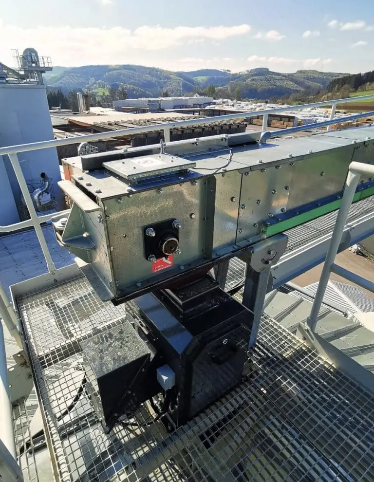 Prallplattenwage auf dem Dach eines Silos, man sieht nur ein quadratisches Rohr mit Messgeräten. Alles steht auf einem Laufgitter, das über eine Treppe erreichbar ist. Im Hintergrund sieht man bewaldete Hügel.