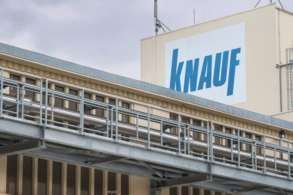 Beiger Industriebau mit großem Logo der Firma Knauf und davor einer Produktionshalle mit Förderrohr.