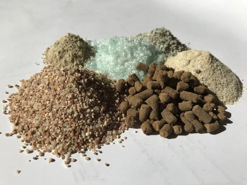 Sechs Häufchen mit Schüttgut, hinten links von dort nach rechts, Sand, Glas, kleine Kiesel, rechts vorn von links nach rechts Linsen große Kiesel, Pressstäbchen in dunkel, Braun und Quarzsand.