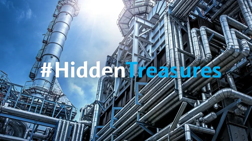Industrieanlage mit sehr vielen Rohren und einem Schornstein vor blauem Himmel. Daraufliegender Text #HiddenTreasures