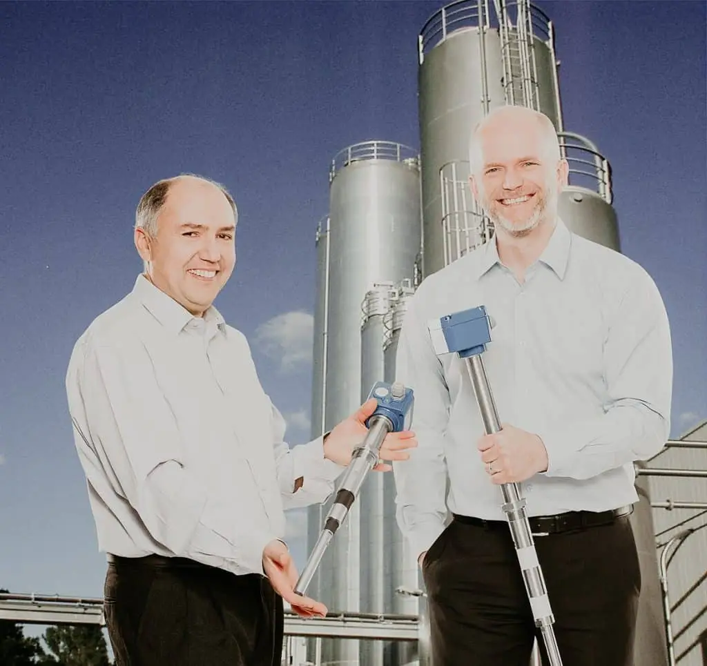 Zwei Ingenieure mit ca. 80 cm langen Grenstandsmessern in der Hand vor einem Silo.