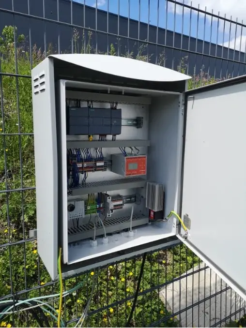Schaltschrank an einem Metallzaun hängend. Er ist geöffnet und man sieht die dort fixierten Elemente und Kabel, alles sehr geordnet.