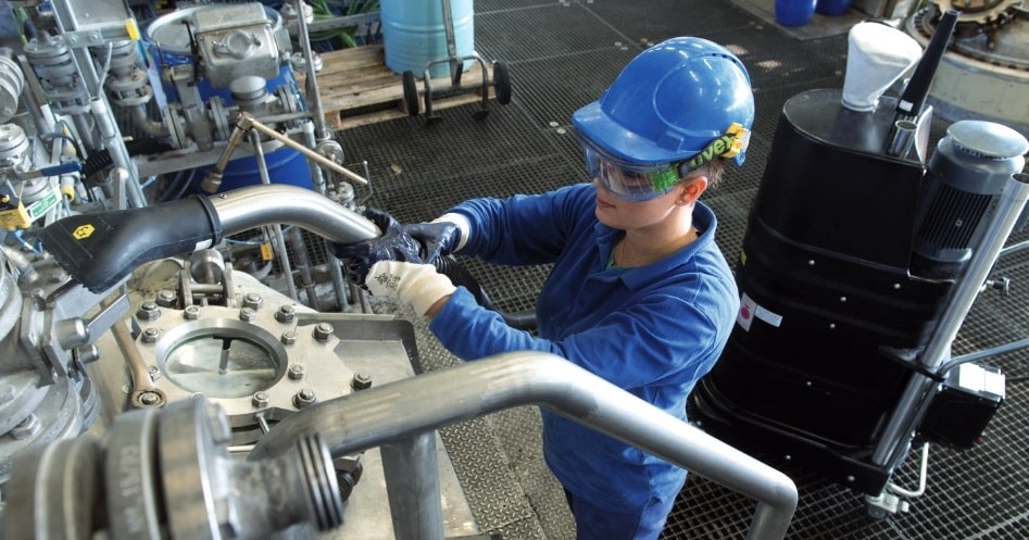 Weibliche Mitarbeiterin mit blauer Arbeitskleidung und Helm saugt eine Industriemaschine ab. Sie hat Handschuhe an, die vorn schwarz und hinten weiß sind.