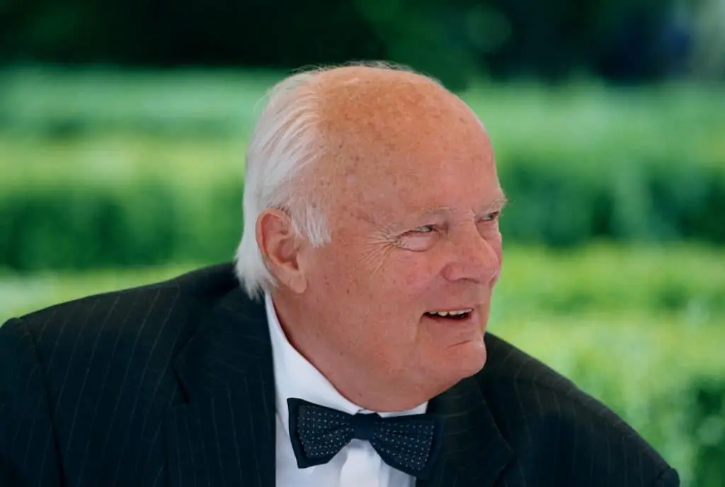 Georg H. Endress in einem grauen Nadelstreifenanzug und mit einer blauen Fliege, deren obere Fliege kleine weiße Sprenkel hat, vor dem verschwommenem Garten-Grün.