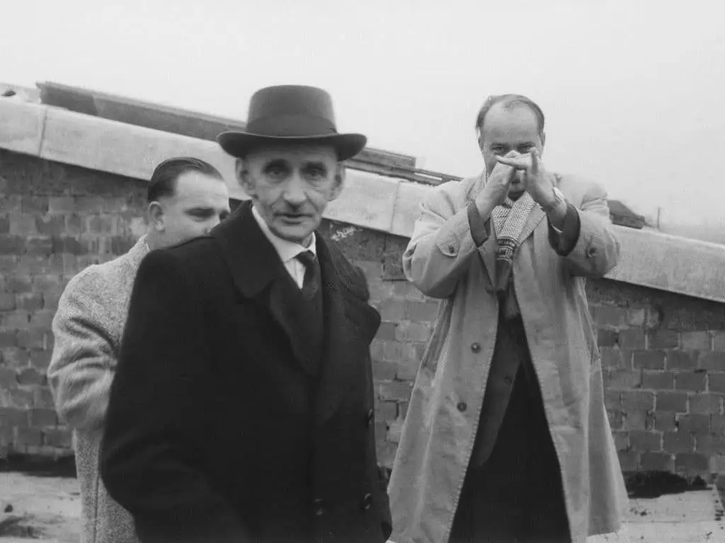 Schwarz weiß Foto von drei Herren, die vor einer abgeschrägten Mauer stehen. Vorn links mit Hut und schwarzem Mandel Ludwig Hauser, hinter ihm ein Herr, der nicht erkennbar ist. Rechts in Regenmantel und Schal, die Hände vorgestreckt, als ob er Trompete spielte, Georg H. Endress.