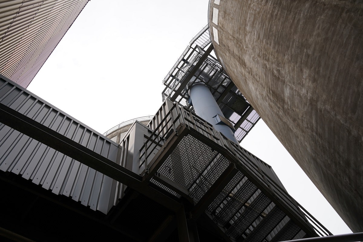 Blick von unten am Vertikalförderer hoch. Links eine Industrie-Stahl-Treppe und eine Industrie-Halle. Rechts ein Turm aus Beton.