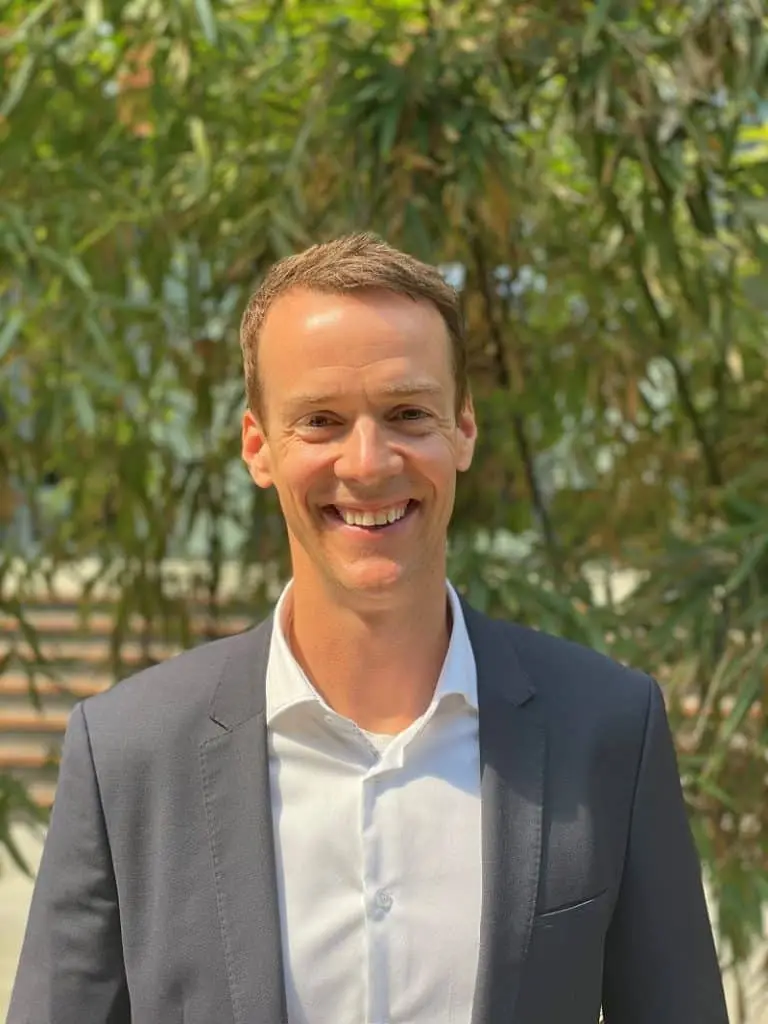 Clemens Reusch lächelt vor Bambus im Anzug ohne Krawatte in die Kamera