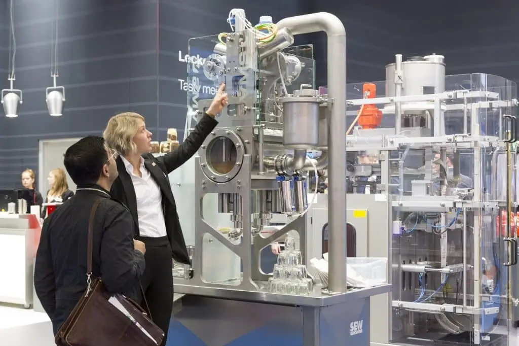 Auf einem Messestand der SEW erklärt eine Mitarbeiterin einem Besucher das Exponat. Links im Hintergrund andere Mitarbeiterinnen am Pult des Messestandes. Weitere Exponate sind zu sehen.