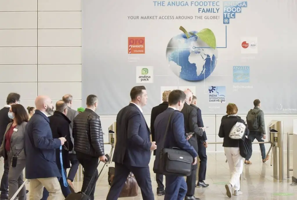 Vor einem großen Plakat der Anuga Foodtec mit einem blauen Apfel auf dem die Kontinente dargestellt sind, gehen Menschen durch Drehkreuze in den Messebereich.