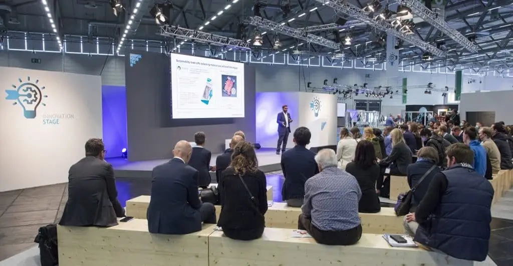 Auf dem Vortragsbereich der Anuga Foodtec sitzen Zuhörer auf Holzboxen und hören interessiert dem Referenten zu, hinter dem auf dem großen Bildschirm eine Präsentation sichtbar ist.