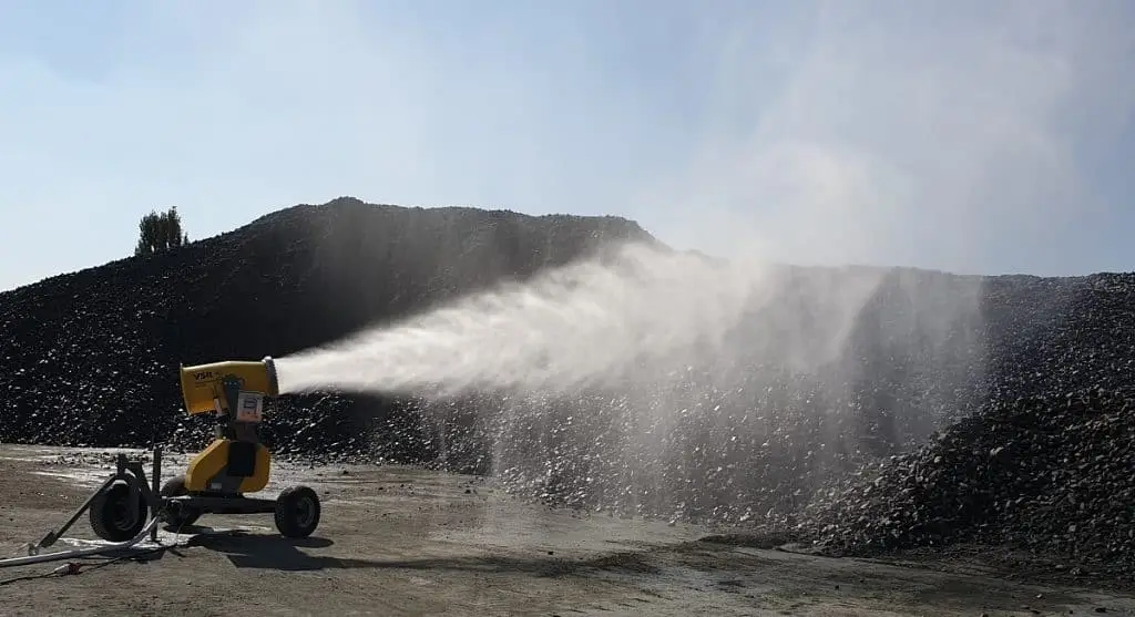 Mobile Staubbindemaschine: Auf einem Außengelände liegt eine Halde mit schwarzem Schüttgut. Davor eine Nebelkanone, die Wasser Richtung Halde versprüht. Die Nebelkanone sieht grundsätzlich aus wie eine Schneekanone. Das Gerät ist gelb und hat das VSR Logo aufgedruckt. Es ist auf drei Rädern aufgesetzt und somit leicht verschiebbar.