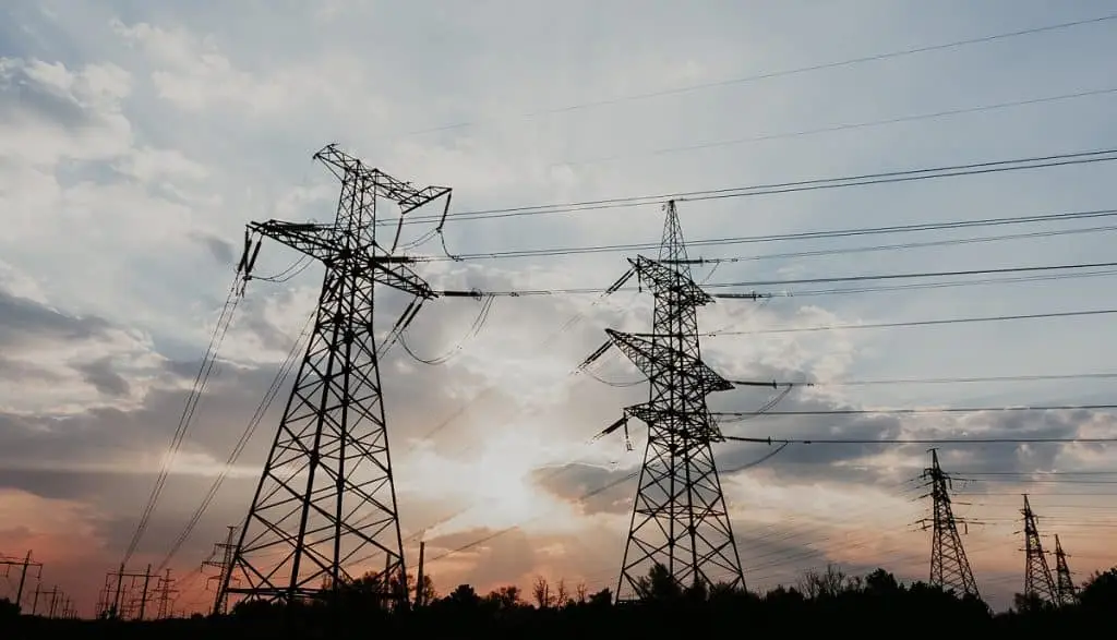 Vor einer untergehenden Sonne mit Wolken davor stehen mehrere Hochspannungsmasten mit Leitungen
