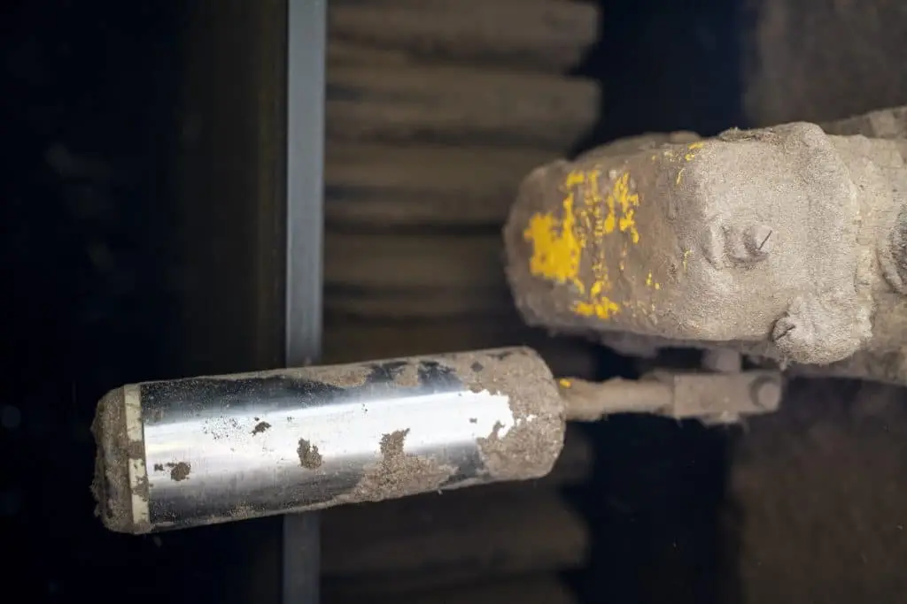 Ein zylindrischer Metallhebel, der voller angebackenem Sand ist. Rechts davon ein offensichtlich gelbes Gerät, das auch voller Sand/Dreck ist.