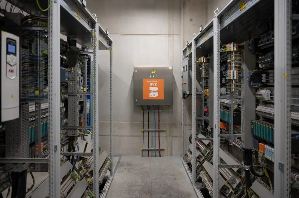 Blick auf eine Wand mit einem Schaltschrank, auf dem ein orangefarbener Aufkleber ist. Rechts und links lauter andere Schalteinheiten, die offen zu sehen sind.
