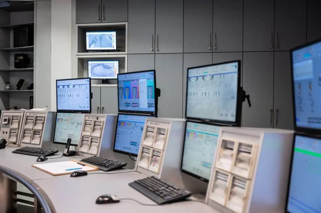 Zentrale mit Bildschirmen, und Messinstrumenten, die alle unterschiedliche Systeme anzeigen. Vor den Bildschirmen Tastaturen, Mäuse und auch ein Telefon.