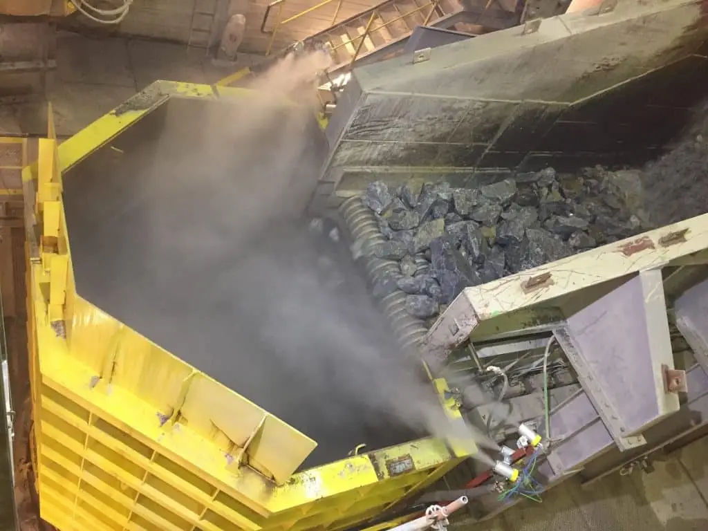 Links ein großer gelber Brecher. Rechts fördert ein Förderband enorme Steine in den Brecher. An der Abwurfstelle versprühen Wasserdüsen feinen Nebel, um den entstehenden Staub zu binden.