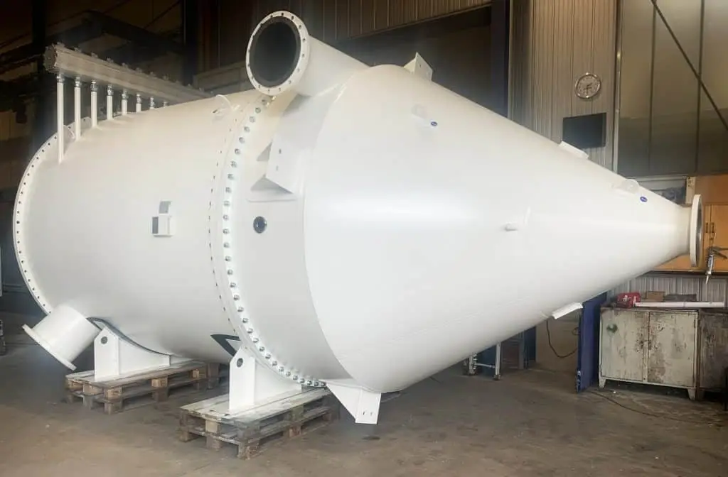 Schlauchfilter Industriegröße. Sieht aus wie eine 3 D Rakete. Die Spitze ist ein Rohrauslauf. Nach der engsten Stelle erweitert sich die Spitze auf Rohrgröße. Das Rohr ist mit Nieten mit der Spitze verbunden. Darüber, rechts, ein Rohreilass. Unten links ein Rohrauslass und hinten auf dem dicken Endrohr sind lauter kleine, dünne Rohre und darüber eine Art metallischer Balken.