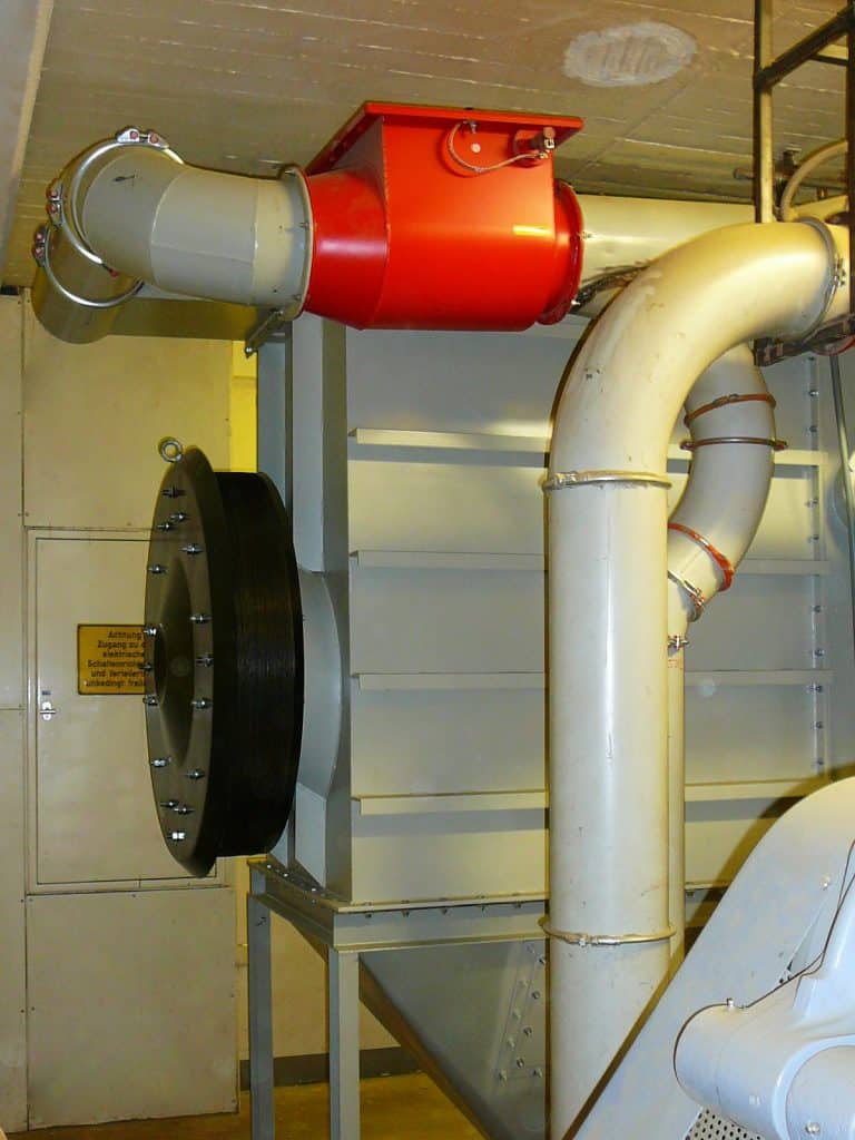 Industrieraum. Links ein metallischer Kubus mit einem schwarzen runden Deckel, der mit Nieten angebracht ist. Darüber und davor Rohre und ein. In dem oberen Rohr ist ein roter Einsatz, der nach oben Trog-artig ausschaut.