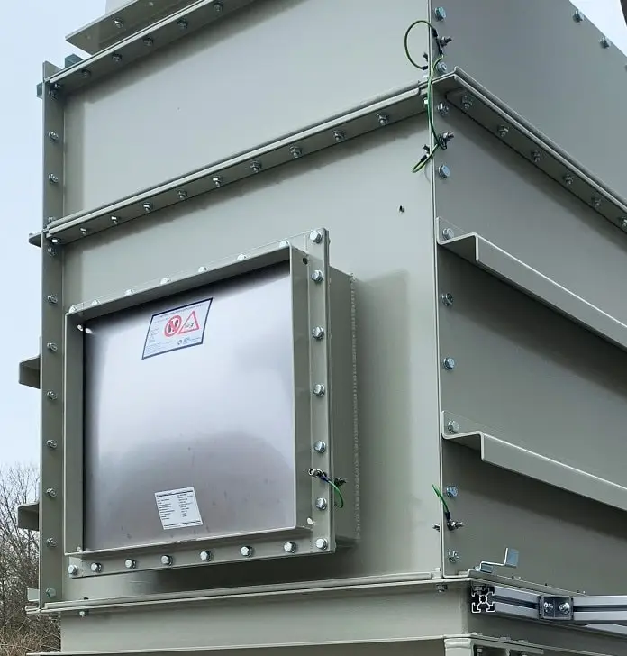Infastaub Taschenfilter, recheckiger Metallkasten mit konischem Auslauf nach unten. Auf der Front ist eine Berstscheibe angebracht. Rohre führen oberhalb und von hinten in den Kubus.